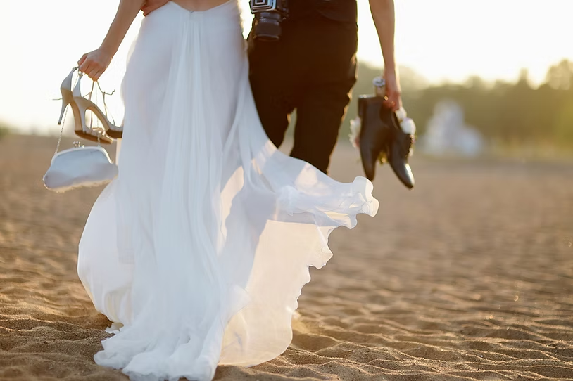 A newlywed couple walks barefoot on the beach, sharing an intimate moment as they carry their shoes and a camera, leaving footprints in the warm sand at sunset.